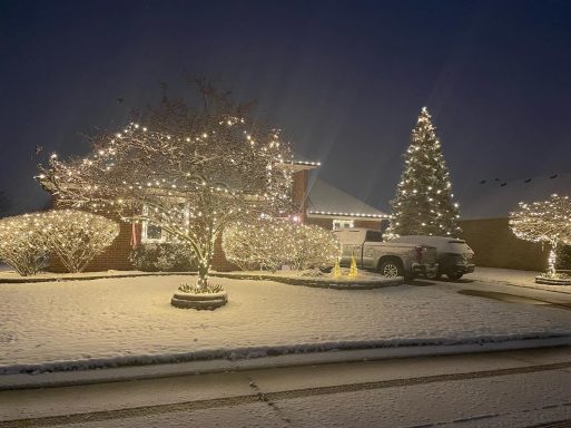 Snow-covered house adorned with festive lights and a decorated Christmas tree at night.